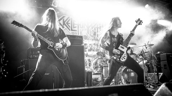 Black-and-white photo of two long-haired guitarists performing on stage with a drummer in the background, surrounded by amplifiers, bright backlighting, and stage smoke during a live metal concert.