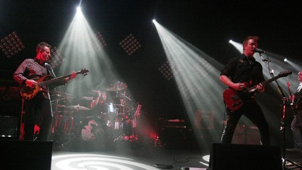 Them Crooked Vultures performing live on stage in 2009, with members playing guitar, bass, and drums under dramatic white spotlights and red stage lighting.