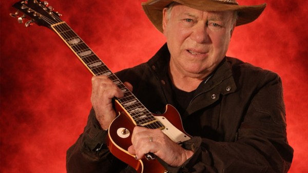 William Shatner holding a Gibson Les Paul guitar, wearing a hat, posed against a bold red background.