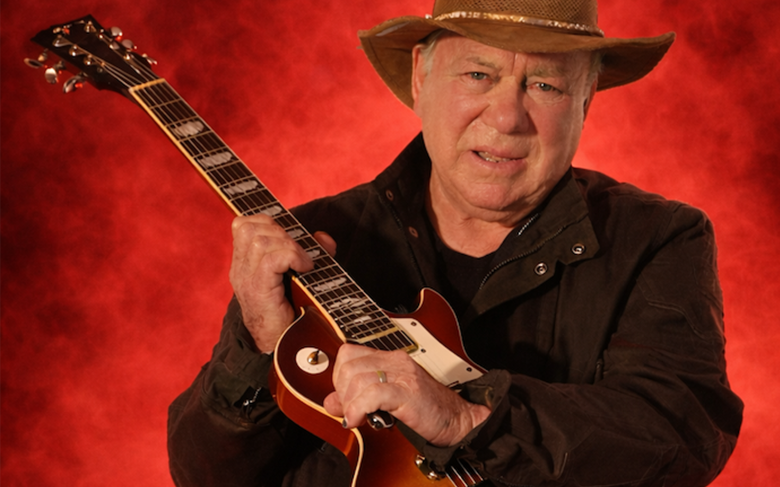 William Shatner holding a Gibson Les Paul guitar, wearing a hat, posed against a bold red background.